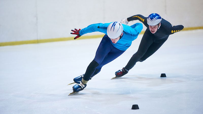 Two male Olympians make a tight turn on an ice skating rink.